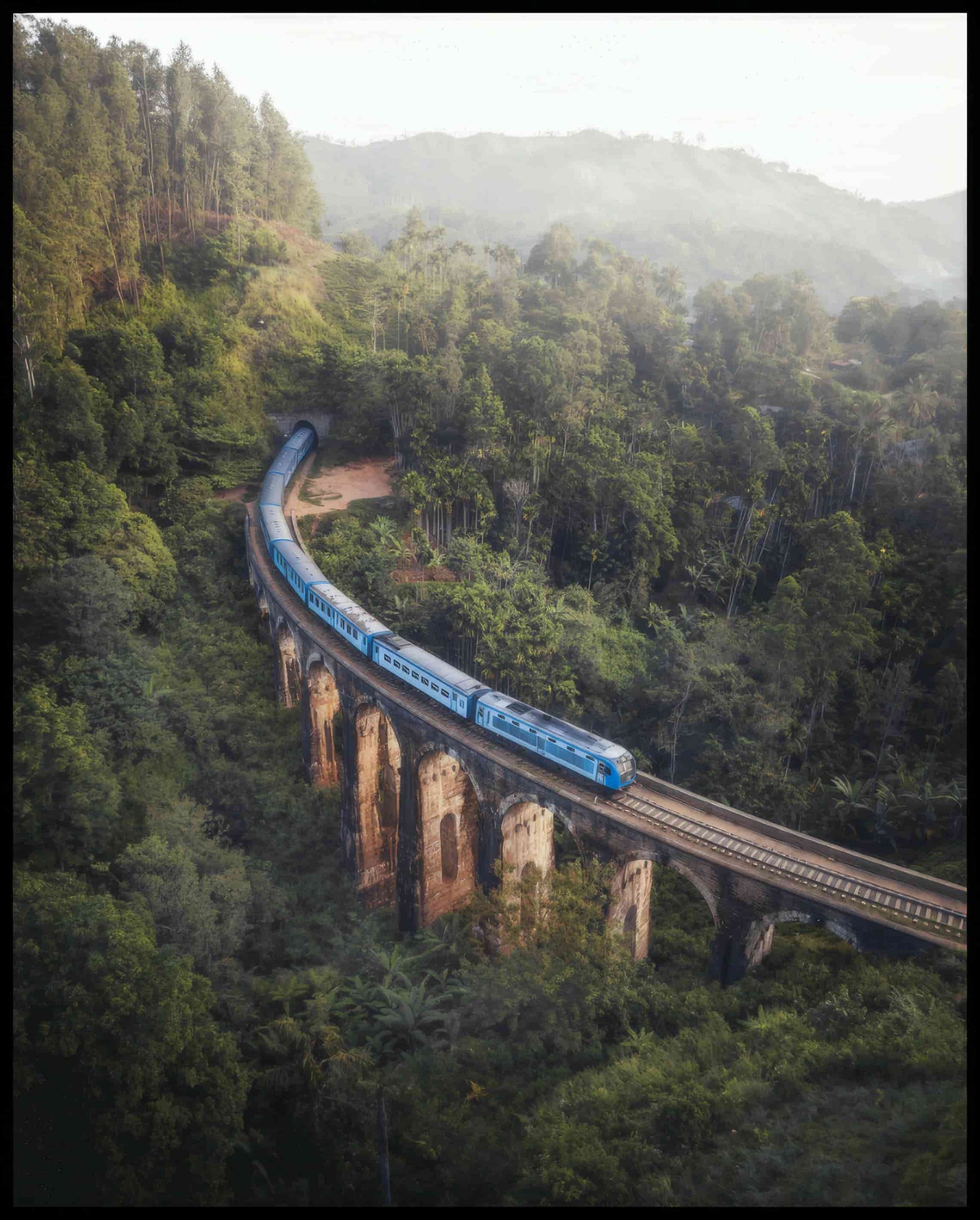Ein Leinwandbild von einem blauen Zug, der über eine alte Brücke inmitten eines grünen Waldes fährt.