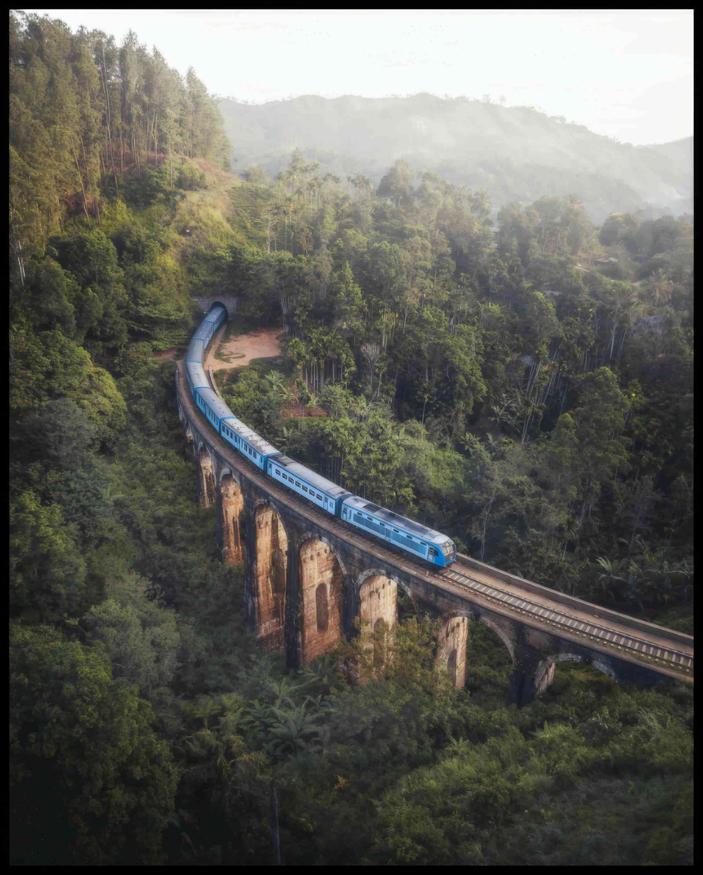 Ein Leinwandbild von einem blauen Zug, der über eine alte Brücke inmitten eines grünen Waldes fährt.