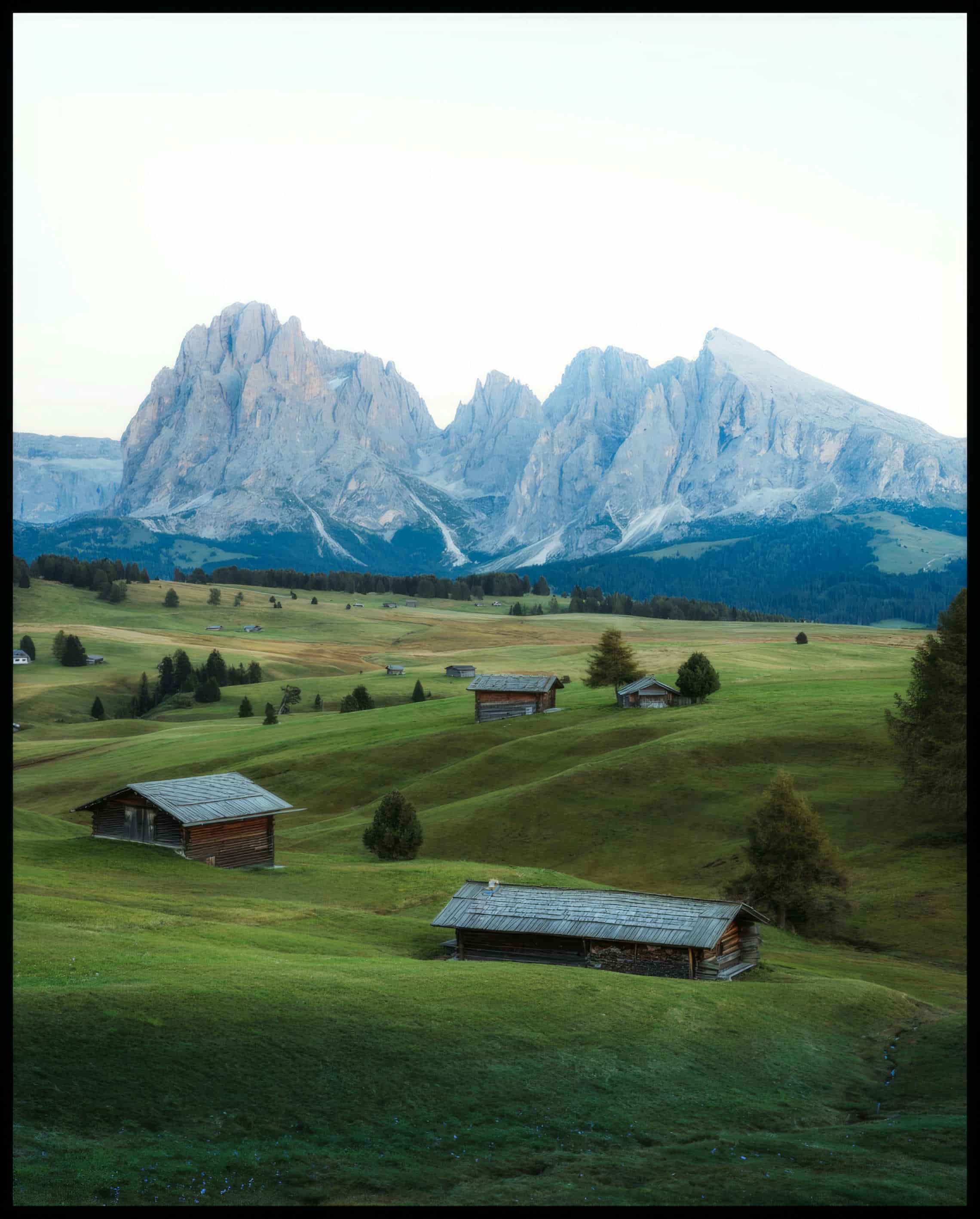 Ein Leinwandbild von einer grünen Alm mit kleinen Hütten und den Dolomiten im Hintergrund.
