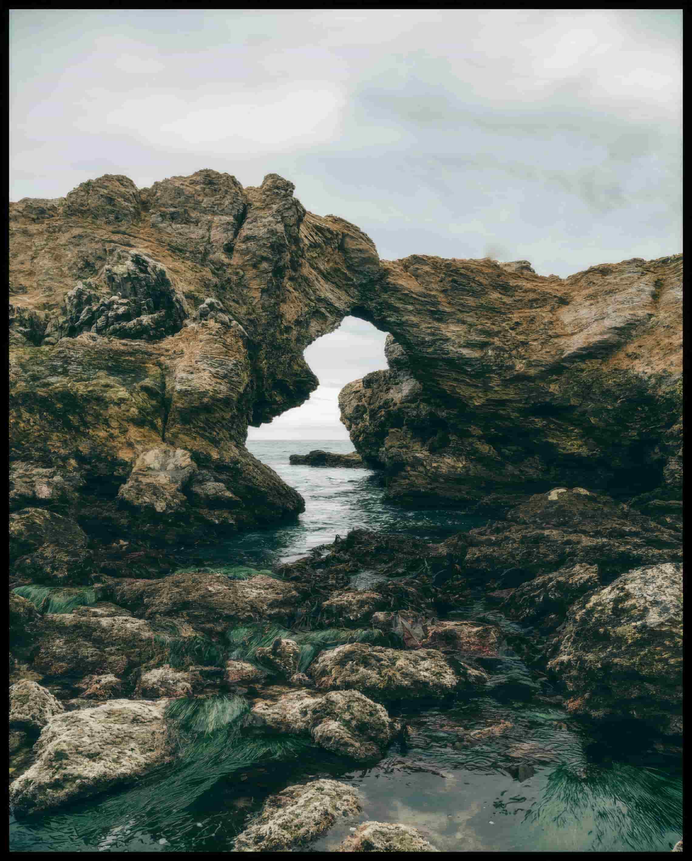 Ein Leinwandbild von einer felsigen Küste mit einer natürlichen Felsformation, die ein Fenster zum Meer bildet.