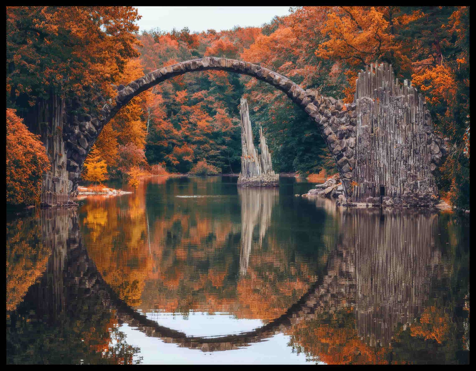 Leinwandbild von der Seitenansicht einer freispannenden Bogenbrücke in einem herbstlichen Wald und über einem ruhigen See.