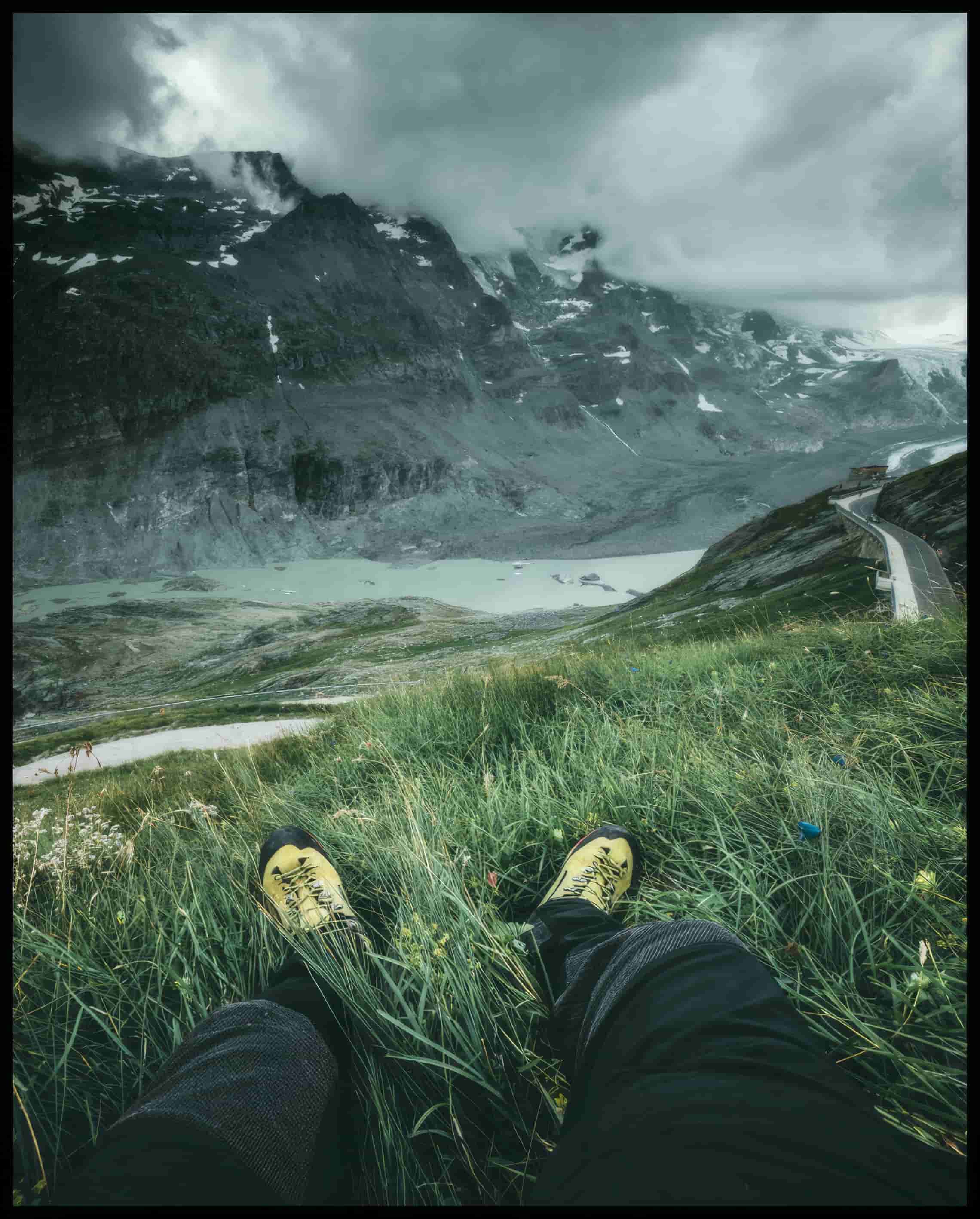 Leinwandbild aus der Egoperspektive von einer Person die im Gras sitzt und auf eine Berglandschaft blickt.