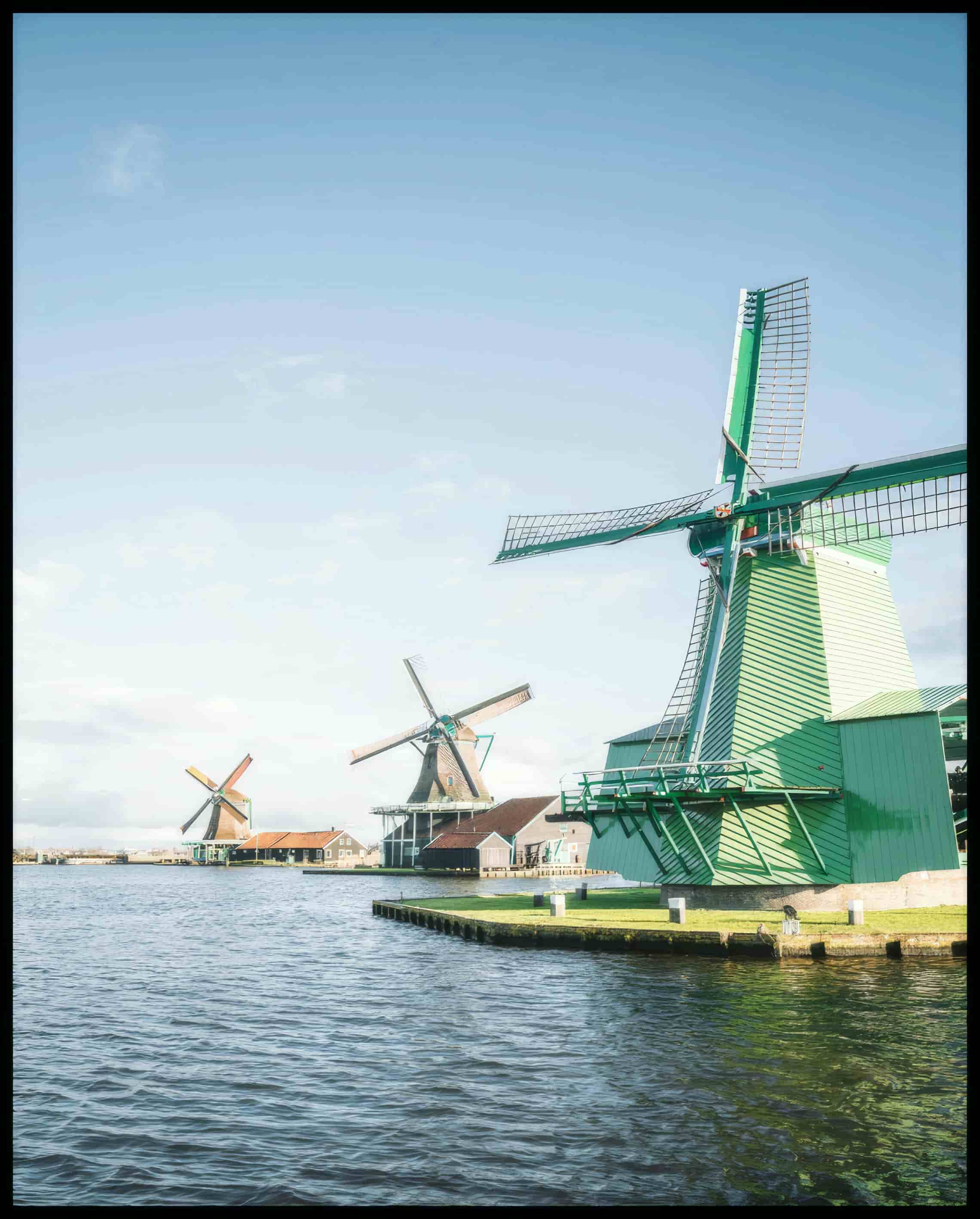 Leinwandbild von drei Windmühlen aus Holz die am Wasser stehen. 