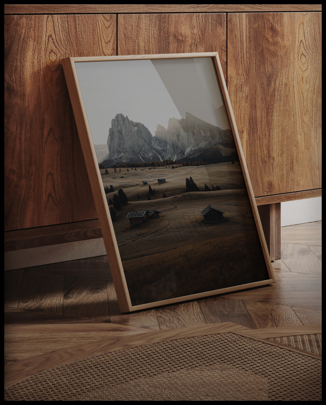 Ein gerahmtes Poster von einer Alm in den Dolomiten bei Sonnenaufgang, an einer Holzkommode angelehnt und stehend auf dem Holzboden