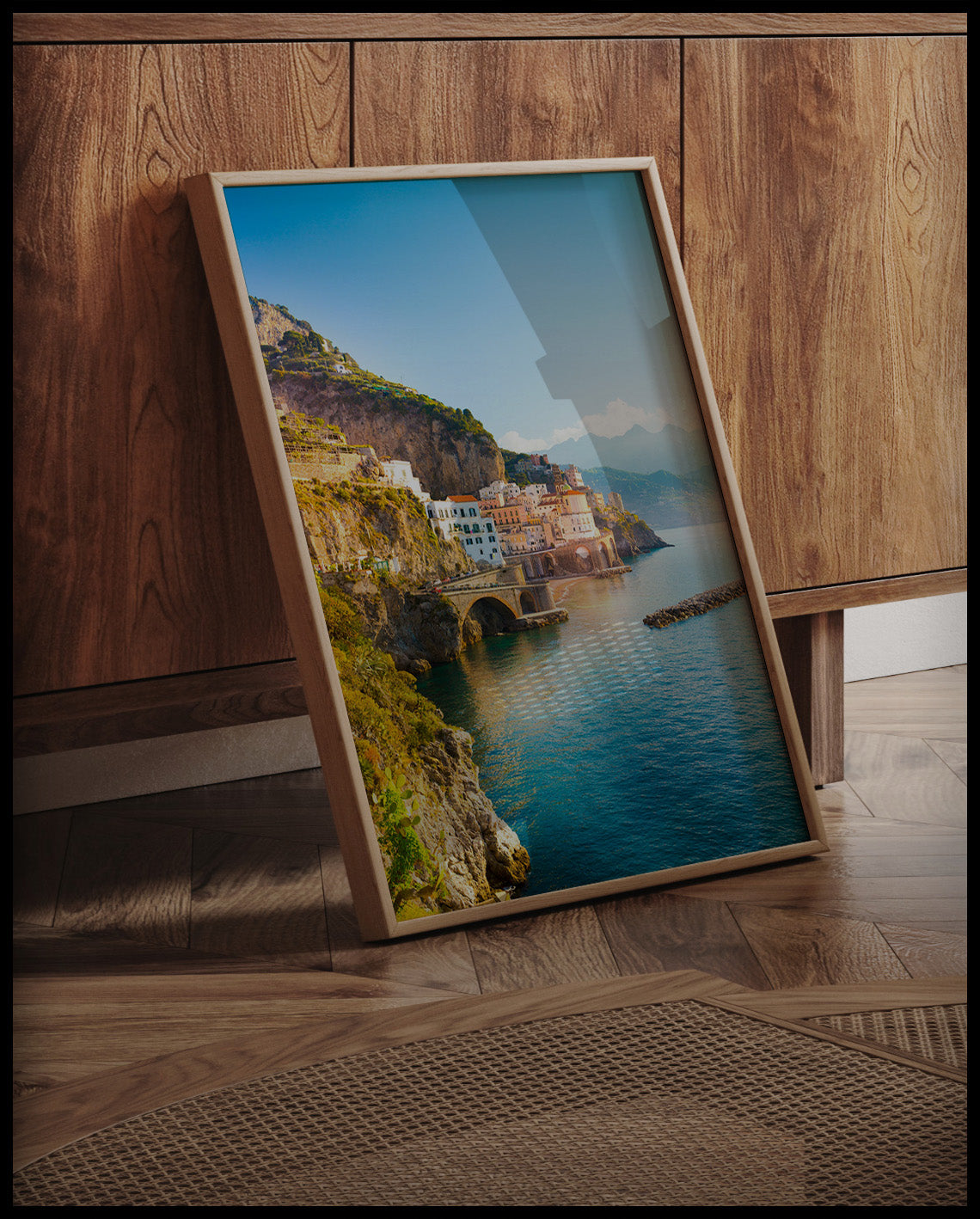Ein gerahmtes Poster von einer Küstenstadt an der Amalfiküste in Italien, mit Häusern, die an steilen Klippen über dem türkisblauen Meer thronen, an einer Holzkommode angelehnt und stehend auf dem Holzboden