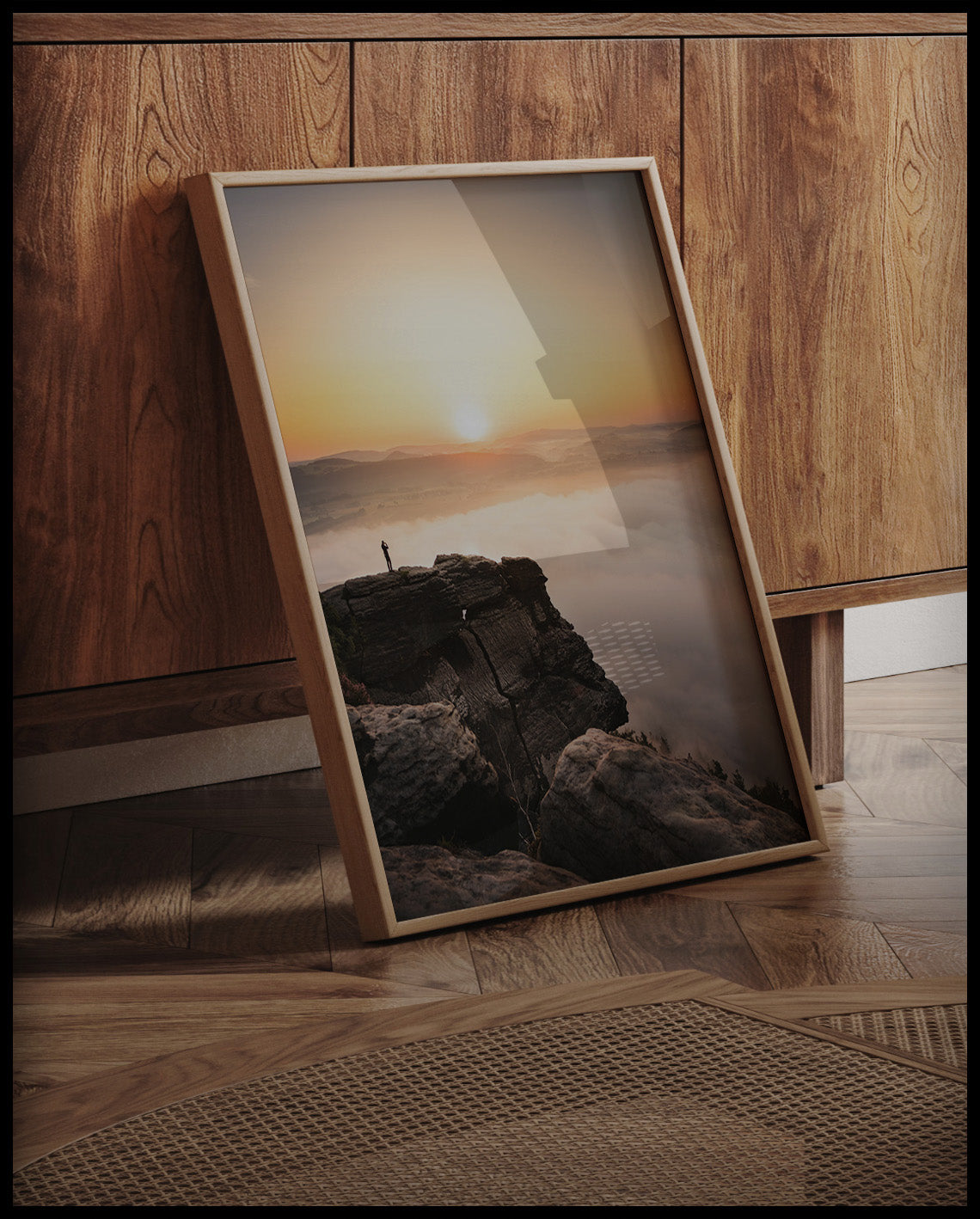 Ein gerahmtes Poster von einem Bergsteiger, der bei Sonnenaufgang auf einer Klippe steht, an einer Holzkommode angelehnt und stehend auf dem Holzboden
