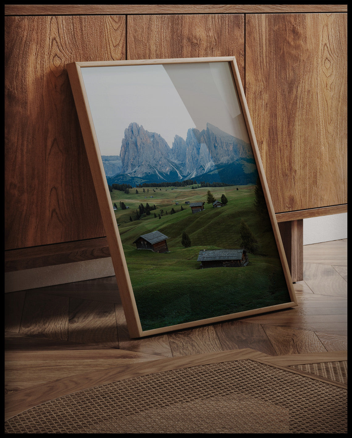 Ein gerahmtes Poster von einer grünen Alm mit kleinen Hütten und den Dolomiten im Hintergrund, an einer Holzkommode angelehnt und stehend auf dem Holzboden