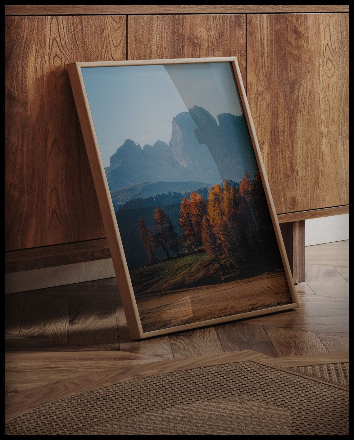 Ein gerahmtes Poster von herbstlich gefärbten Bäumen vor einer Berglandschaft in den Dolomiten, an einer Holzkommode angelehnt und stehend auf dem Holzboden