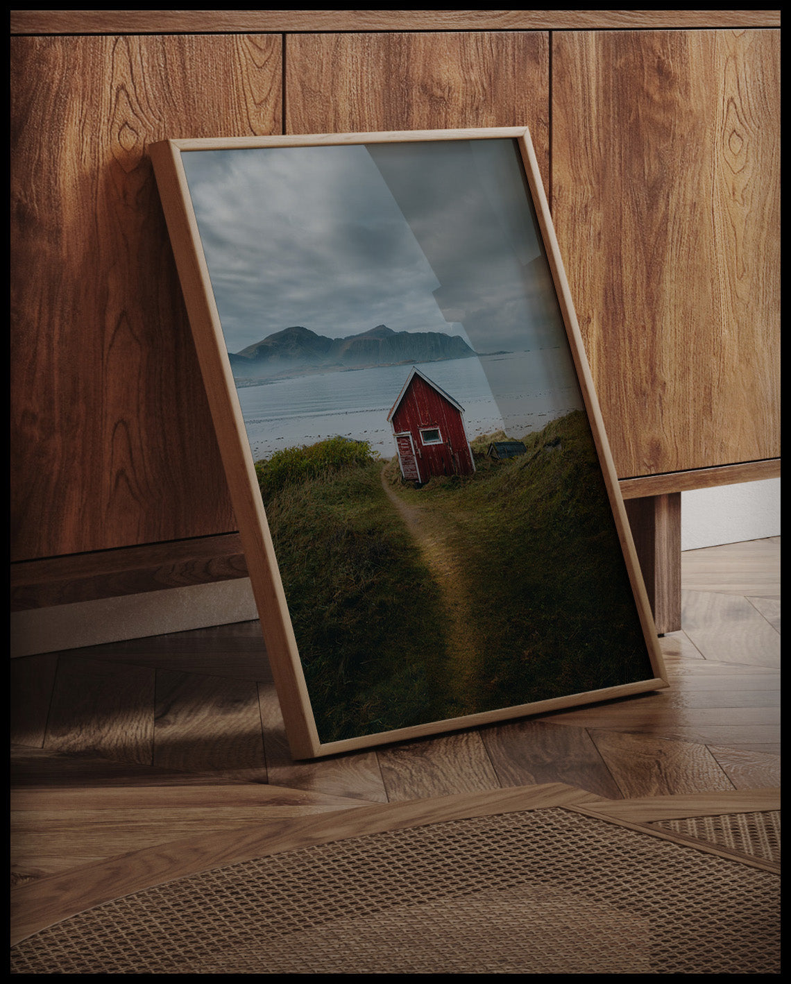 Ein gerahmtes Poster von einem kleinen roten Haus am Seeufer mit Bergen im Hintergrund, an einer Holzkommode angelehnt und stehend auf dem Holzboden