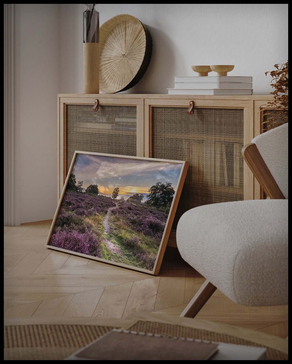 Ein gerahmtes Poster von einer Heidefläche mit lila Blumen im Sonnenuntergang, an einer Holzkommode angelehnt und stehend auf dem Holzboden
