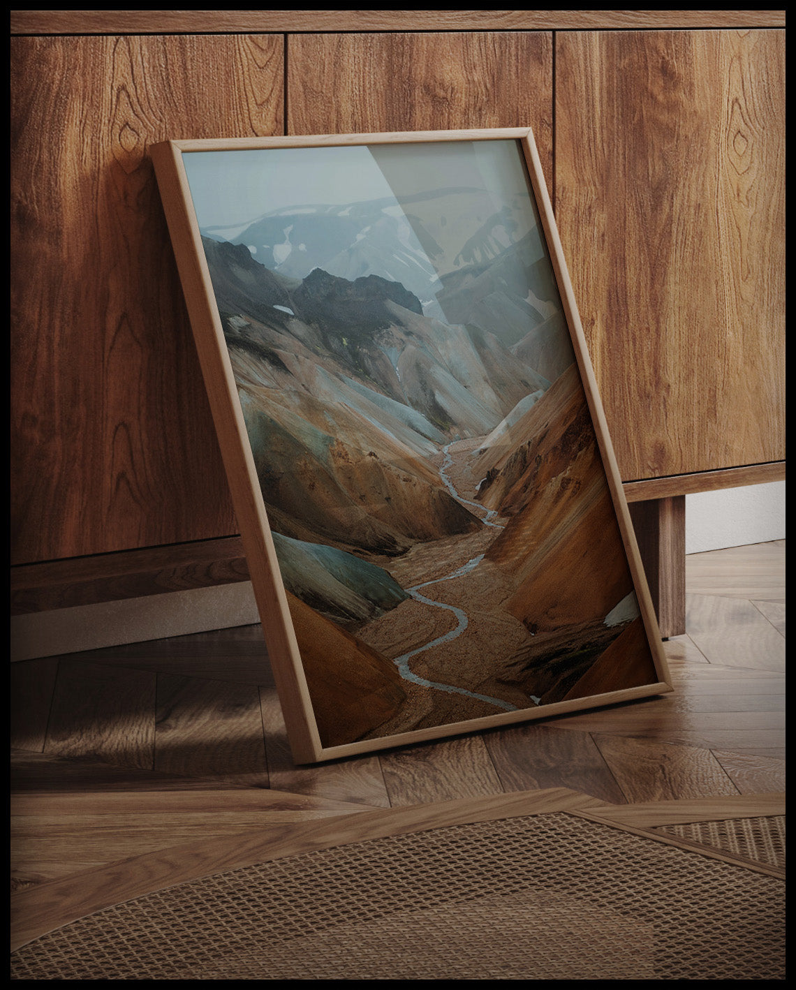 Ein gerahmtes Poster von einem Tal mit einem Fluss und orangenen Bergen, an einer Holzkommode angelehnt und stehend auf dem Holzboden