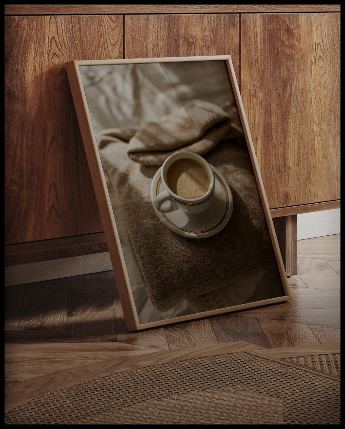 Ein gerahmtes Poster von einer Tasse Kaffee auf einem gemütlichen, beigen Pullover, an einer Holzkommode angelehnt und stehend auf dem Holzboden