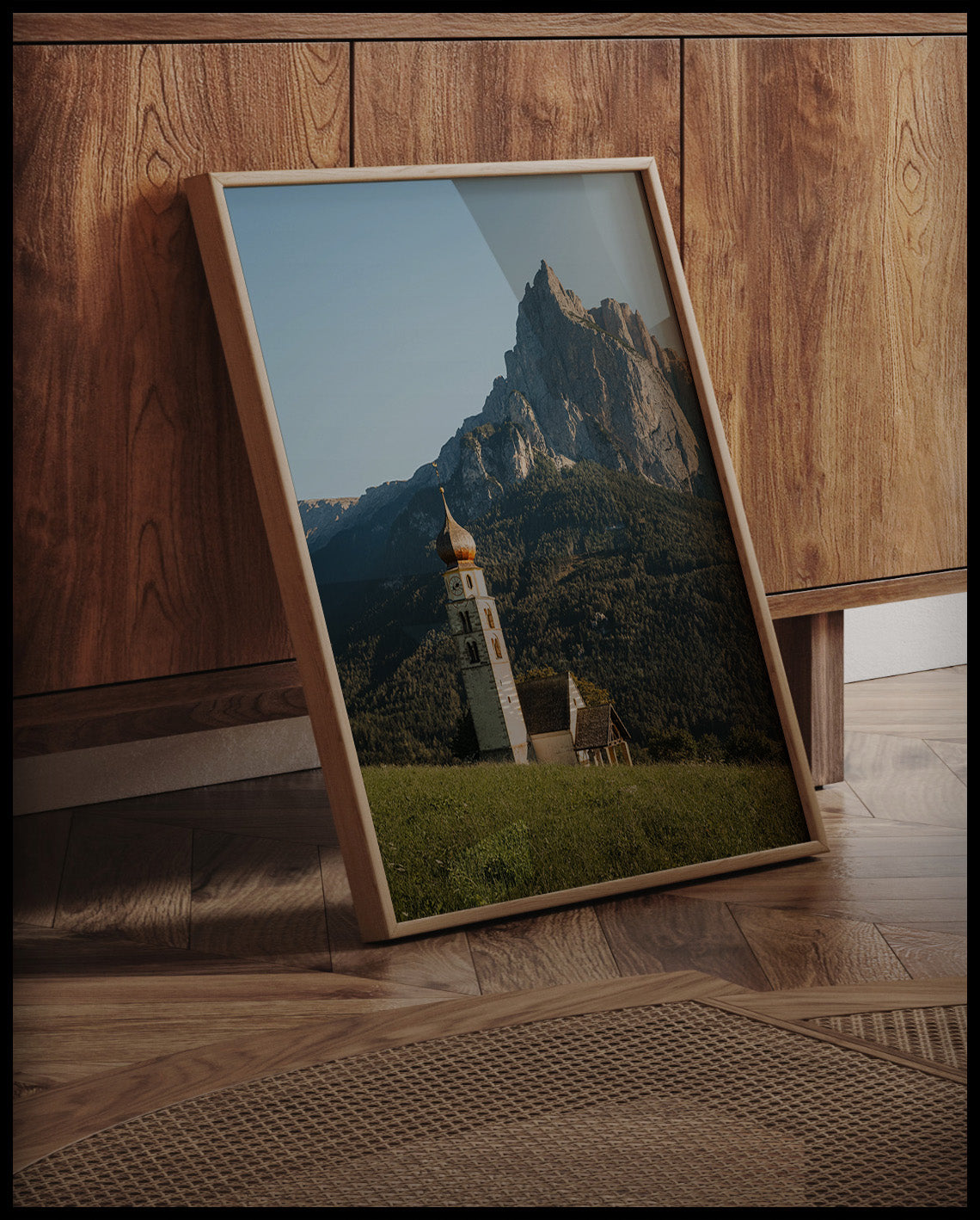 Ein gerahmtes Poster von einer Kirche am Abhang eines Berges in den Dolomiten, an einer Holzkommode angelehnt und stehend auf dem Holzboden