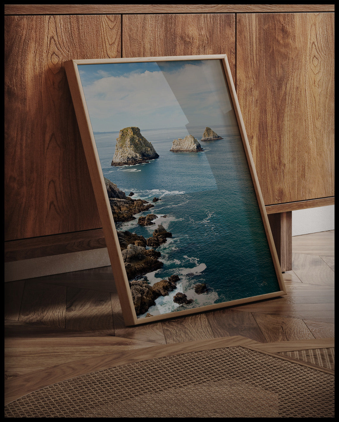 Ein gerahmtes Poster vom Blick auf das blaue Meer und mehrere Felsformationen die aus dem Wasser blicken, an einer Holzkommode angelehnt und stehend auf dem Holzboden