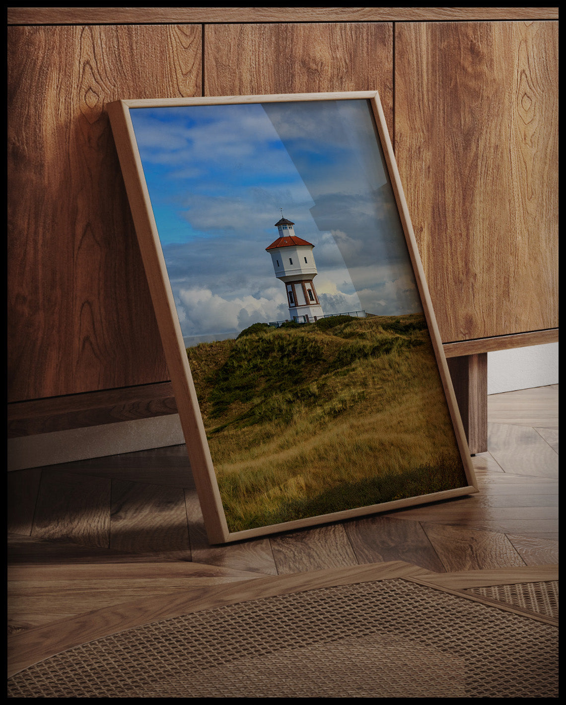Ein gerahmtes Poster von einem weißen Wasserturm auf der Insel Langeoog, an einer Holzkommode angelehnt und stehend auf dem Holzboden