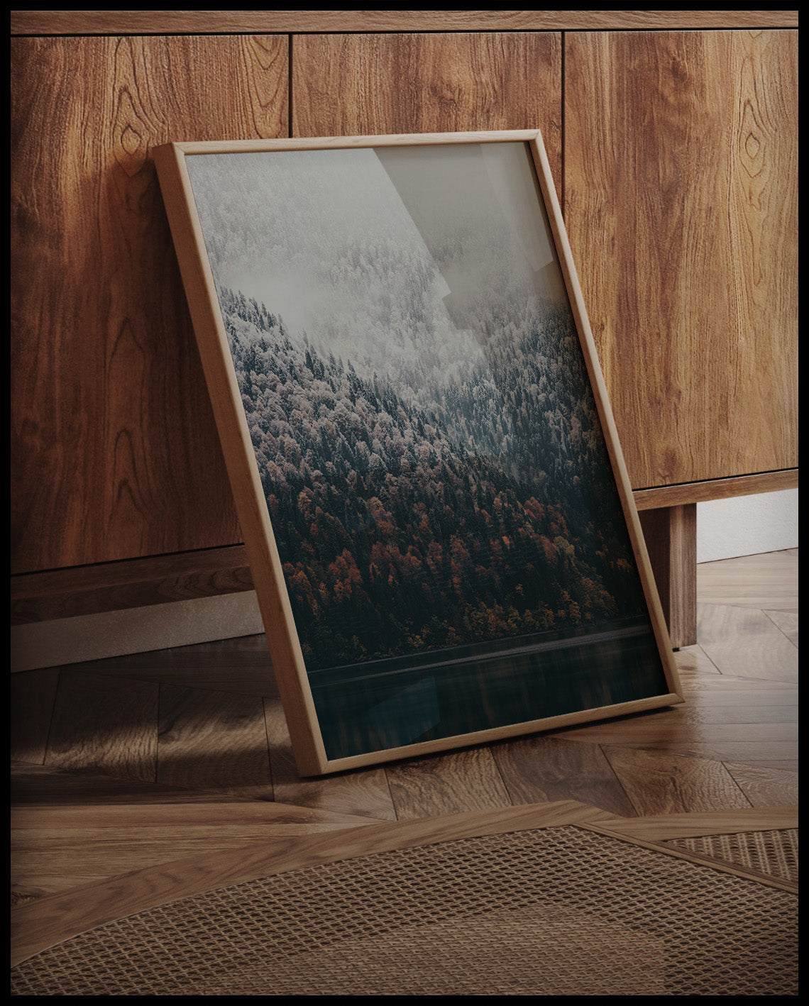 Ein gerahmtes Poster von einem nebeligen Nadelwald, der stellenweise von Schnee bedeckt ist und an einen See grenzt, an einer Holzkommode angelehnt und stehend auf dem Holzboden
