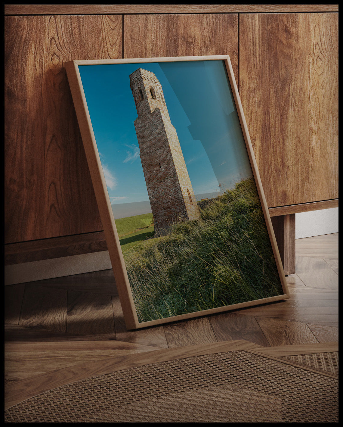 Ein gerahmtes Poster von einem steinernen Turm auf einer grünen, hügeligen Wiese, an einer Holzkommode angelehnt und stehend auf dem Holzboden