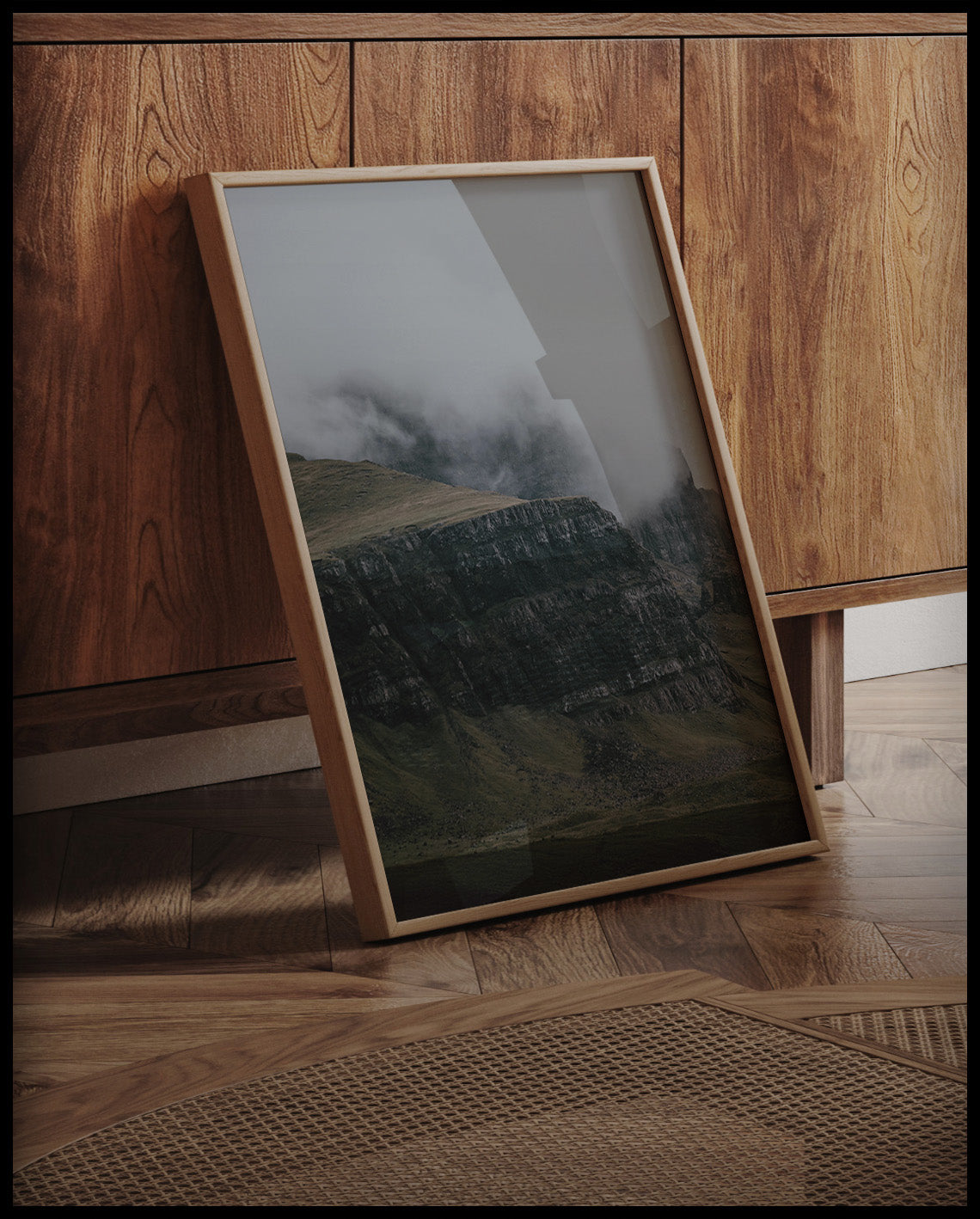 Ein gerahmtes Poster von einem steilen Abhang eines Berges, der von Gras bedeckt und von Nebel umschlungen ist, an einer Holzkommode angelehnt und stehend auf dem Holzboden