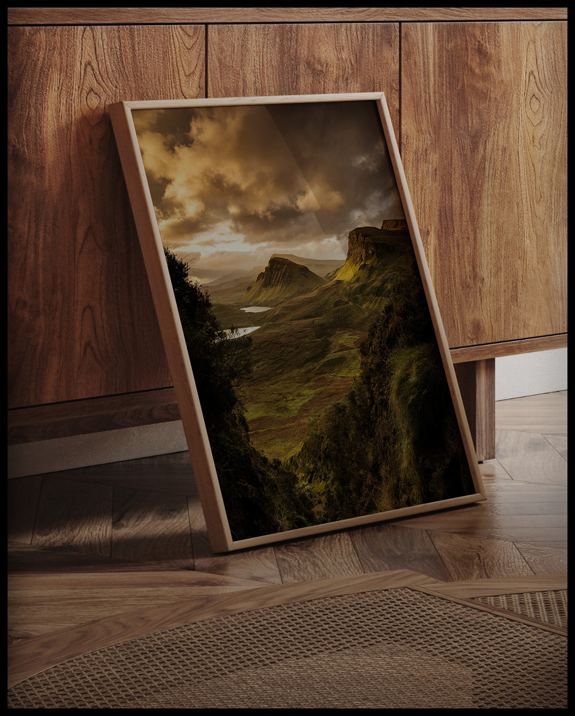 Ein gerahmtes Poster von einer kargen, grünen Berglandschaft im Sonnenuntergang, an einer Holzkommode angelehnt und stehend auf dem Holzboden