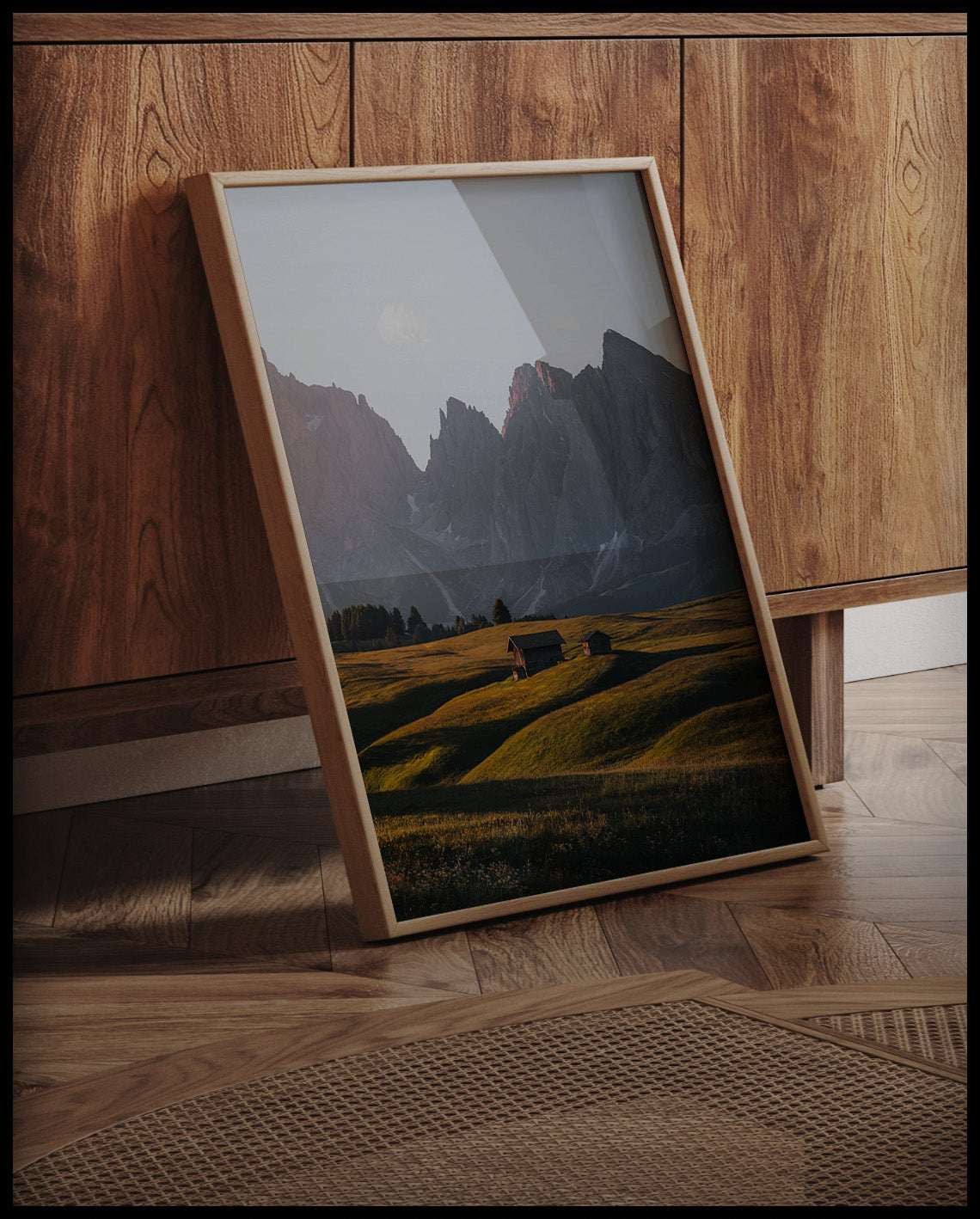 Ein gerahmtes Poster von zwei Holzhütten auf einer goldenen Wiese vor einer steilen, kargen Berglandschaft, an einer Holzkommode angelehnt und stehend auf dem Holzboden