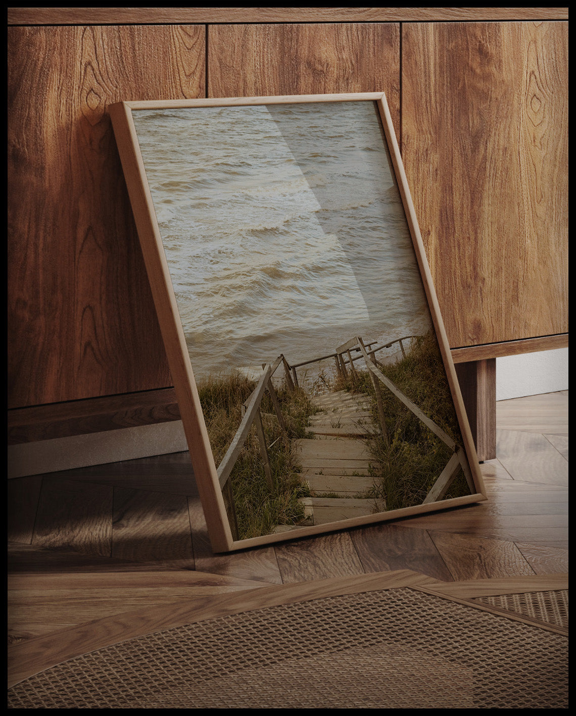 Ein gerahmtes Poster von einer Holztreppe die durch Dünen runter zum Strand führt, an einer Holzkommode angelehnt und stehend auf dem Holzboden
