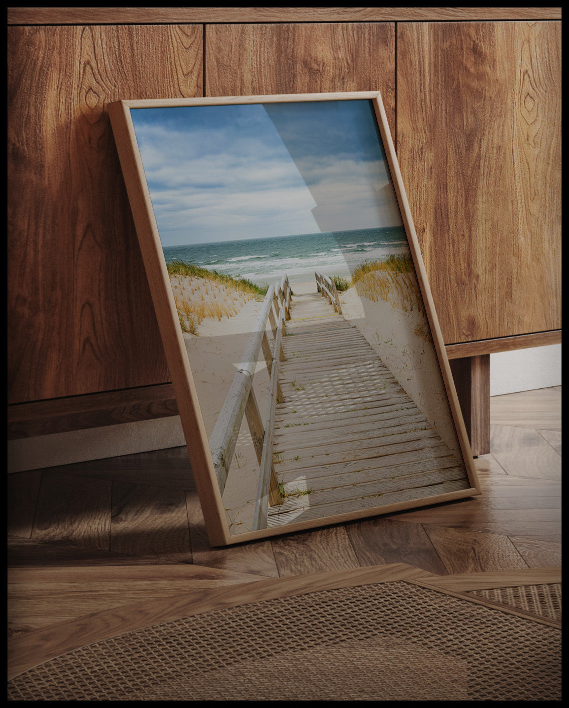 Ein gerahmtes Poster von einem Steg aus Holz der durch sandige Dünen zum Strand führt, an einer Holzkommode angelehnt und stehend auf dem Holzboden
