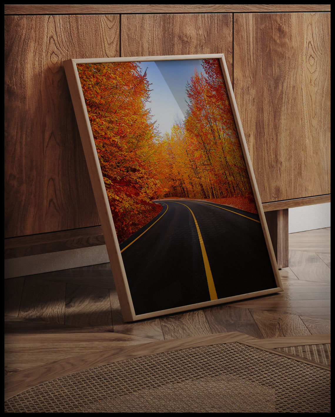 Ein gerahmtes Poster von einer leeren schwarzen Straße die durch einen herbstlichen. orangenen Wald führt, an einer Holzkommode angelehnt und stehend auf dem Holzboden