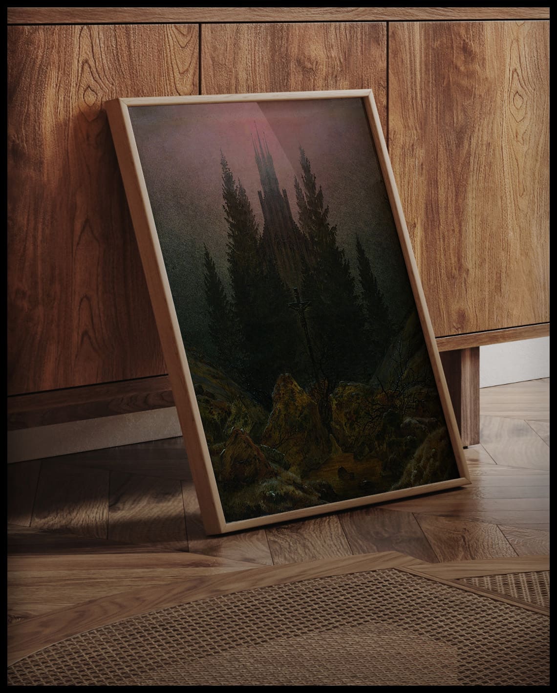 Ein gerahmter Kunstdruck mit einem Kreuz in einer felsigen Landschaft, dahinter eine Kirche umgeben von Tannen, an eine Holzkommode angelehnt und stehend auf dem Holzboden