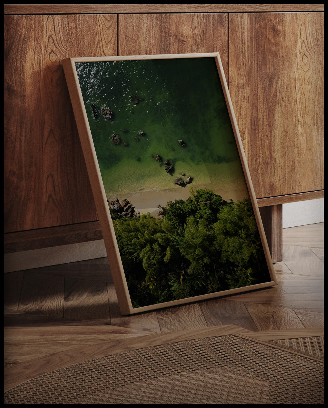 Ein gerahmtes Poster von einem Sandstrand mit grünlichem Wasser vor einem tropischen Wald, an einer Holzkommode angelehnt und stehend auf dem Holzboden