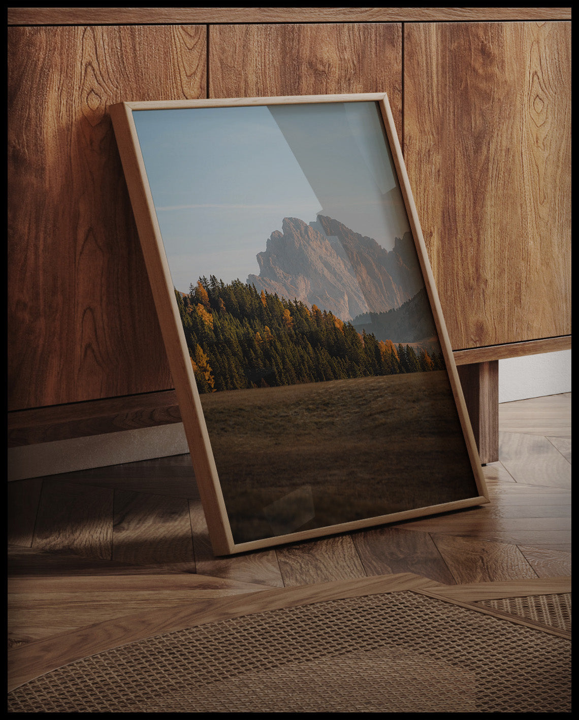 Ein gerahmtes Poster von einem dichten Waldstück in einer kargen Berglandschaft, an einer Holzkommode angelehnt und stehend auf dem Holzboden
