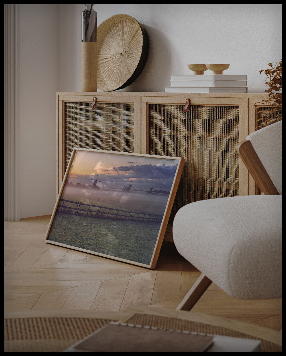 Ein gerahmtes Poster von einer nebligen Wiese und einem Zaun, hinter denen sich Windmühlen verbergen, an einer Holzkommode angelehnt und stehend auf dem Holzboden