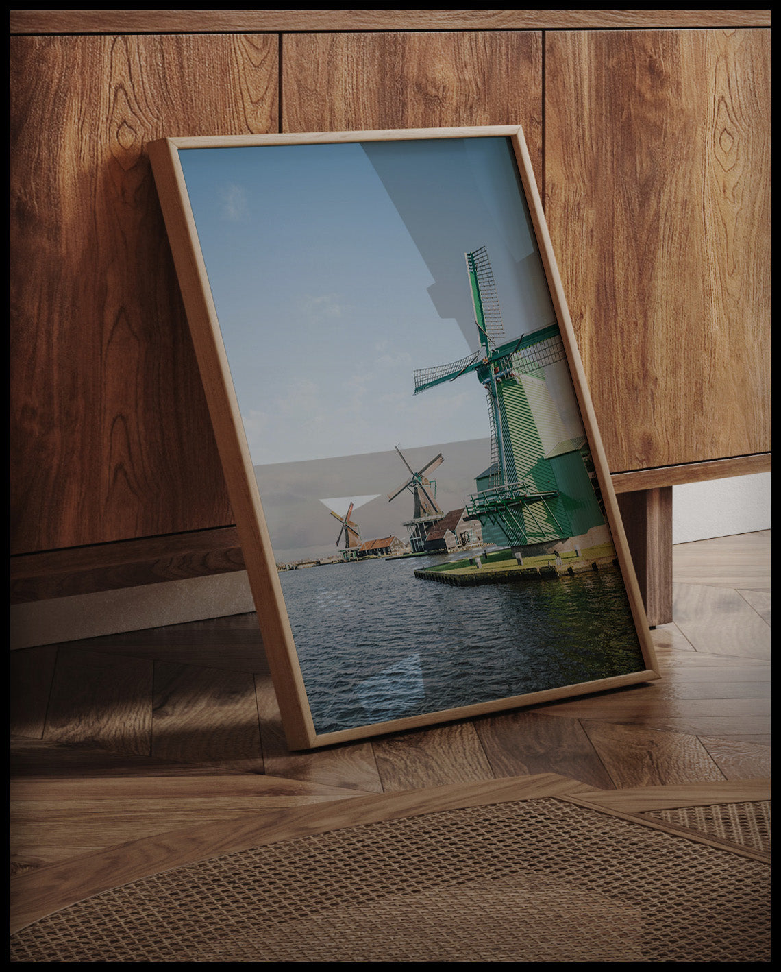Ein gerahmtes Poster von drei Windmühlen aus Holz die am Wasser stehen, an einer Holzkommode angelehnt und stehend auf dem Holzboden
