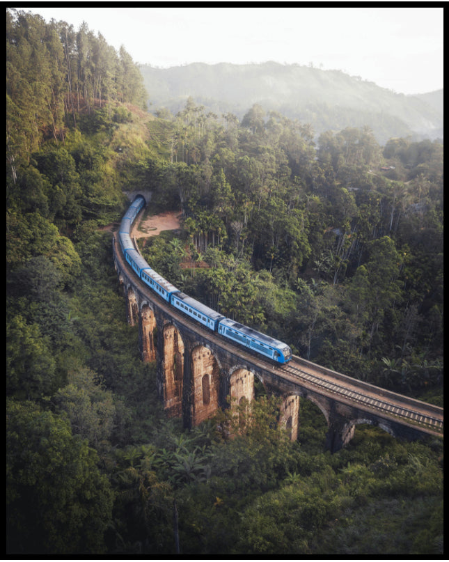 Ein Poster von einem blauen Zug, der über eine alte Brücke inmitten eines grünen Waldes fährt.