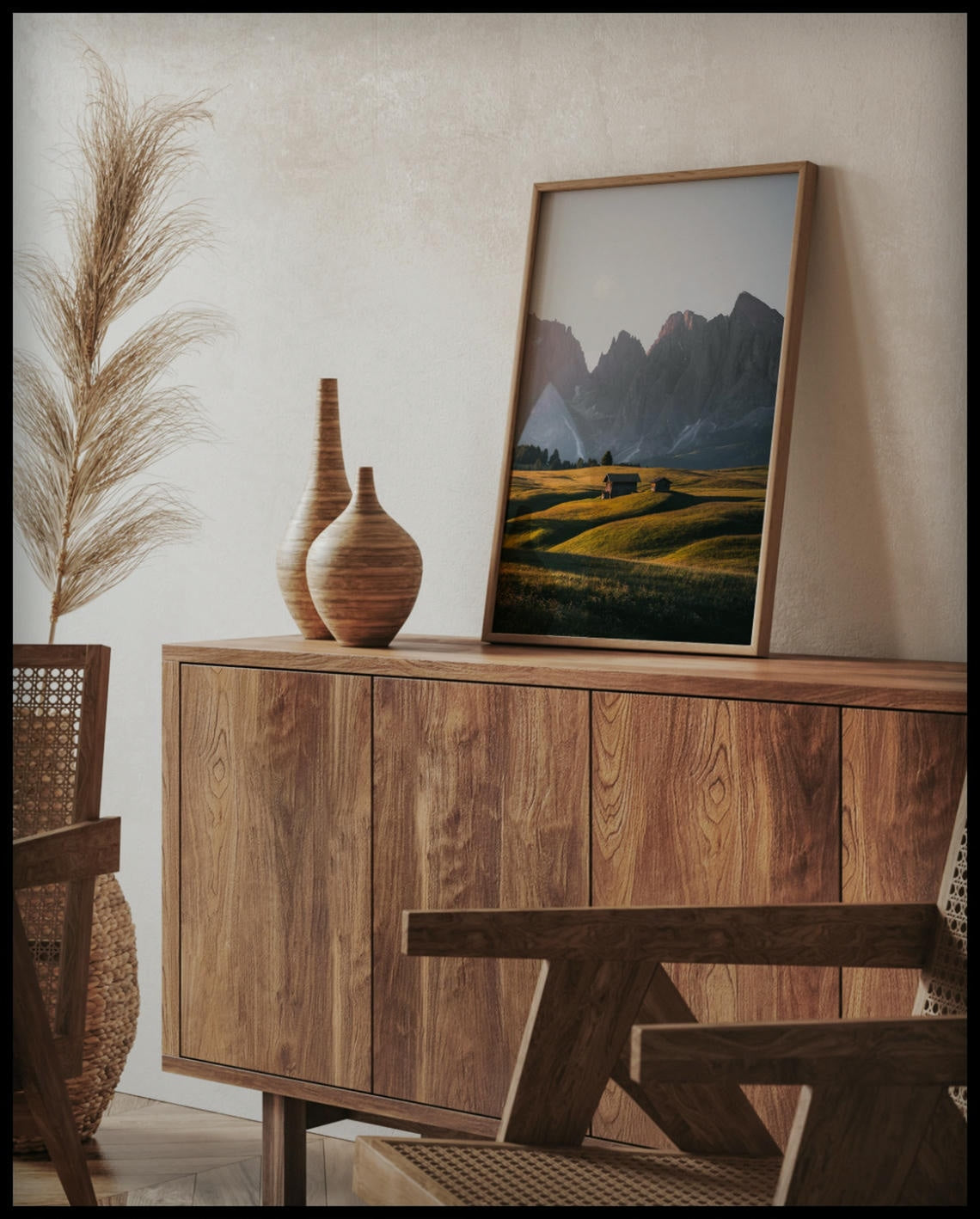 Ein gerahmtes Poster von zwei Holzhütten auf einer goldenen Wiese vor einer steilen, kargen Berglandschaft, auf einer Holzkommode an die Wand angelehnt