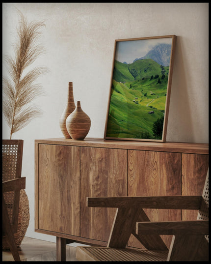 Ein gerahmtes Poster von einer grünen Berglandschaft mit einzelnen Bäumen und Holzhütten, auf einer Holzkommode an die Wand angelehnt