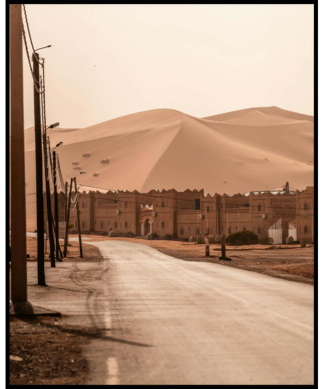 Ein Poster von einer Straße, die zu einer Siedlung am Rande einer weitläufigen Wüstenlandschaft mit hohen Sanddünen führt.