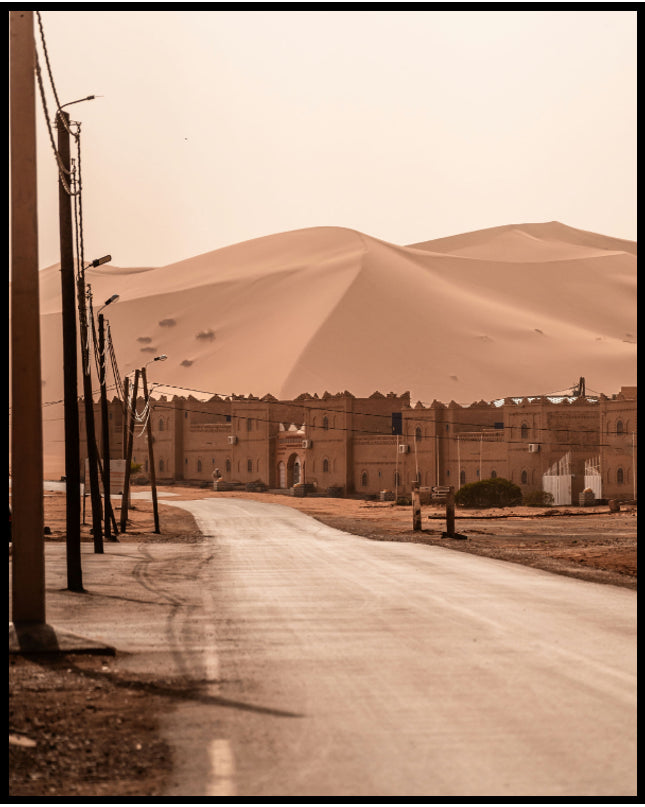 Ein Poster von einer Straße, die zu einer traditionellen Siedlung am Rande einer weitläufigen Wüstenlandschaft mit hohen Sanddünen führt.