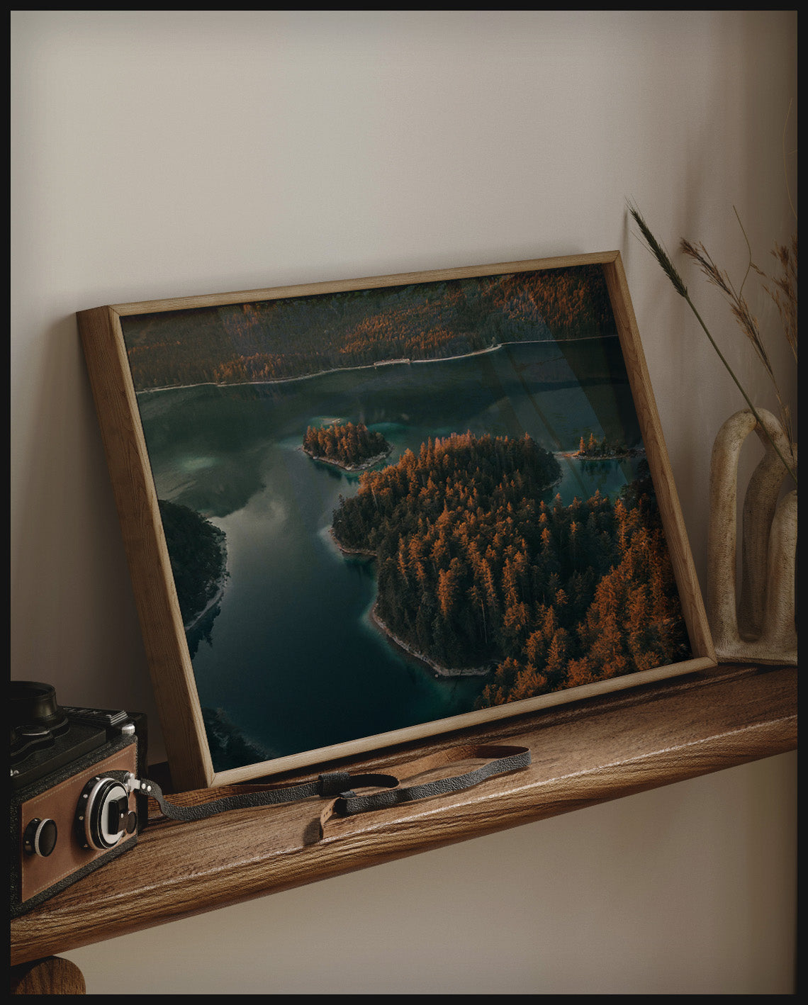 Ein gerahmtes Poster von einer Insel in einem ruhigen See, umgeben von Bäumen, auf einem Holzregal an die Wand angelehnt