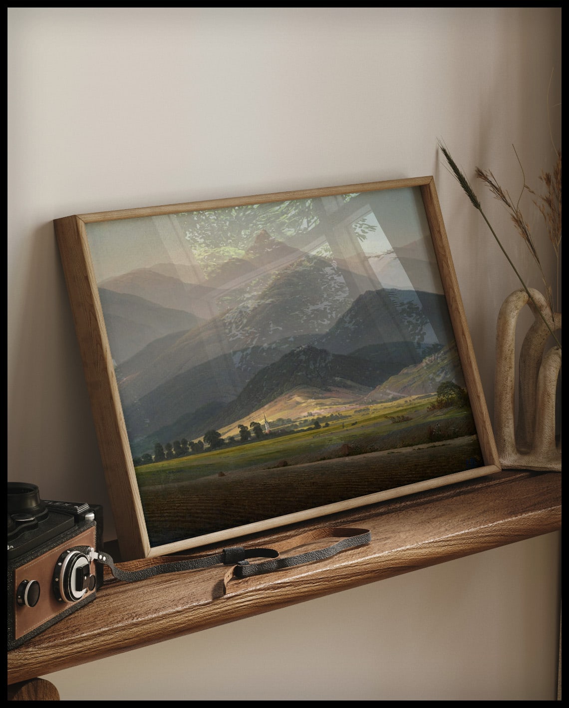 Ein gerahmter Kunstdruck von einer weitläufigen Berglandschaft mit vielen Äckern und Wiesen im Vordergrund, auf einem Holzregal an die Wand angelehnt