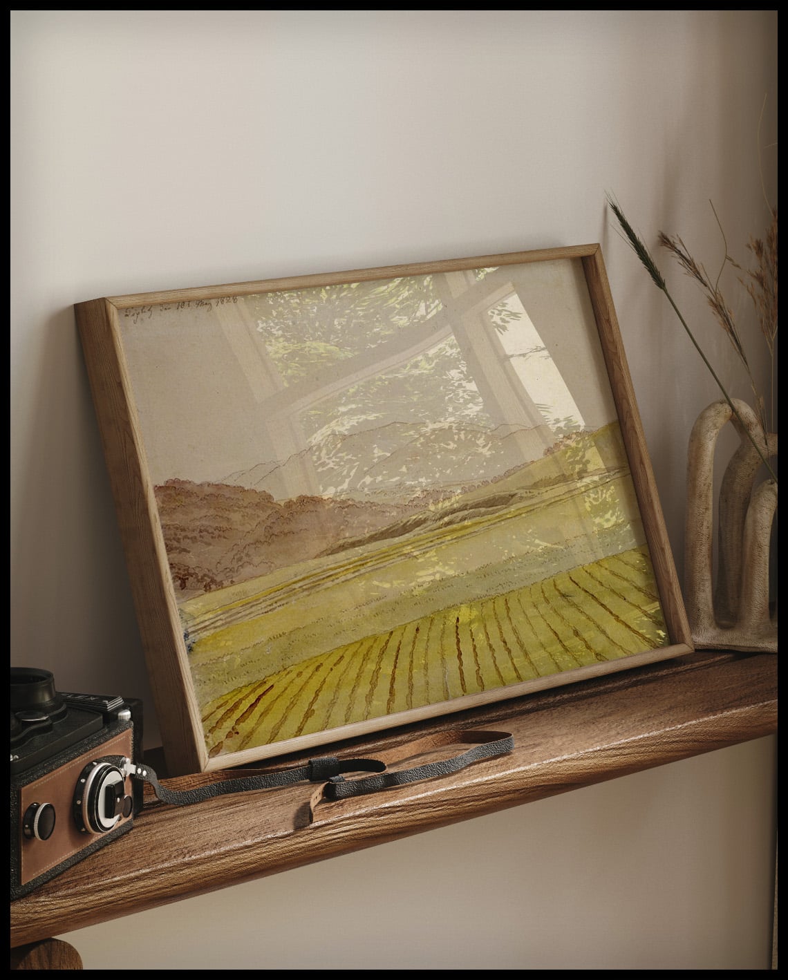 Ein gerahmter Kunstdruck von einem Feld und bewaldeten Bergen im Hintergrund, auf einem Holzregal an die Wand angelehnt