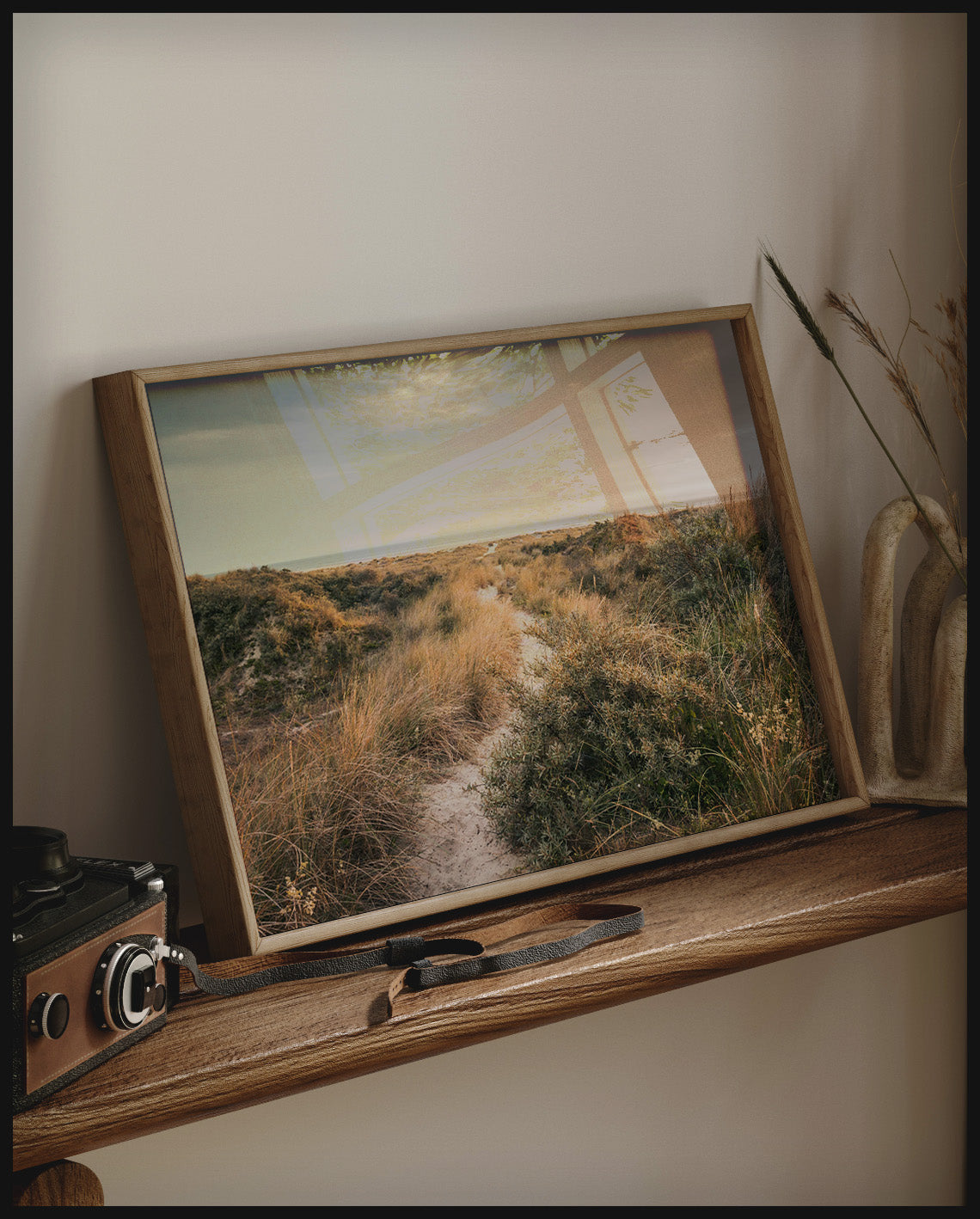 Ein gerahmtes Poster von einer Wanderung durch grasige Dünen, auf einem Holzregal an die Wand angelehnt