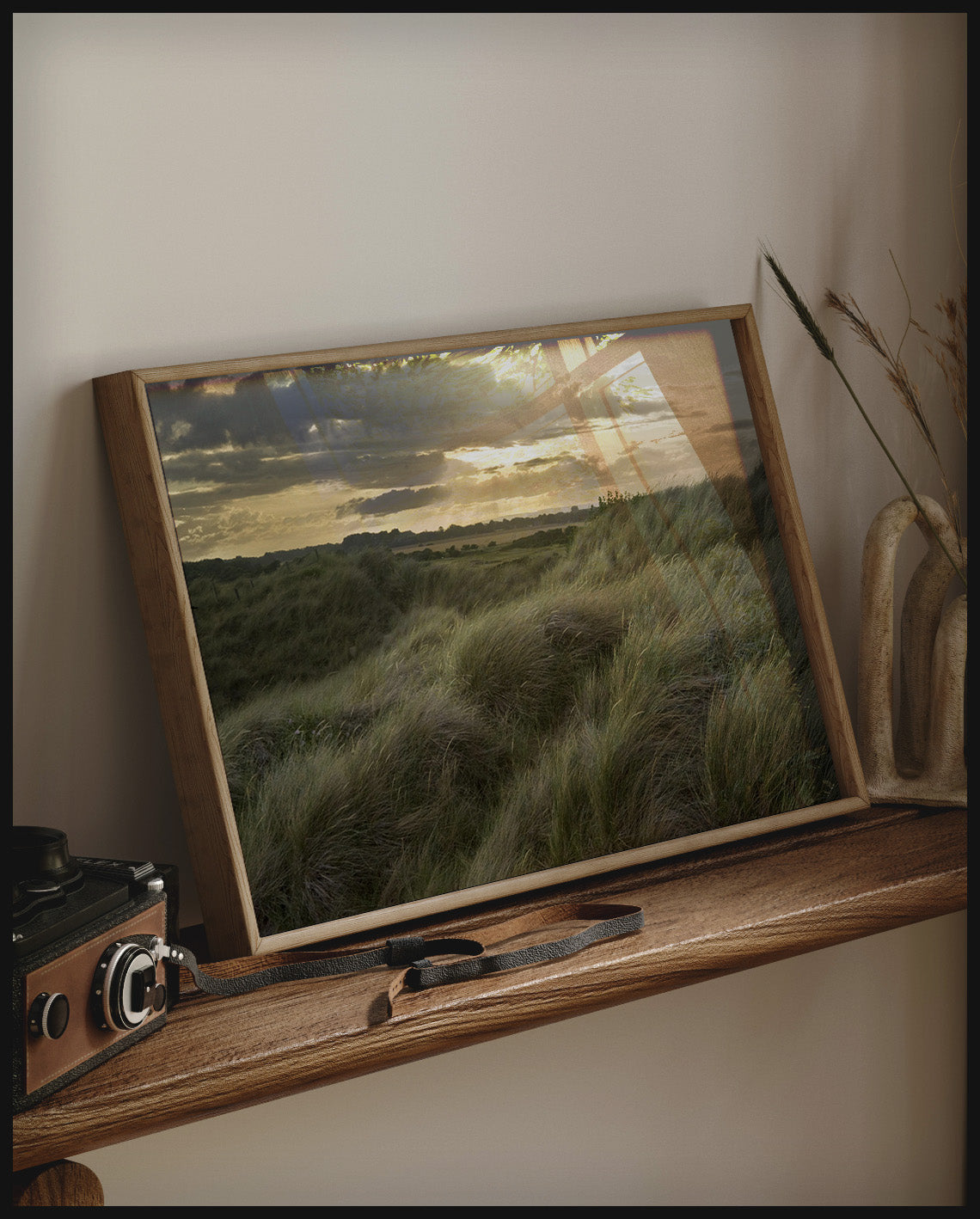 Ein gerahmtes Poster von Gräsern auf einer Düne bei Sonnenuntergang, auf einem Holzregal an die Wand angelehnt