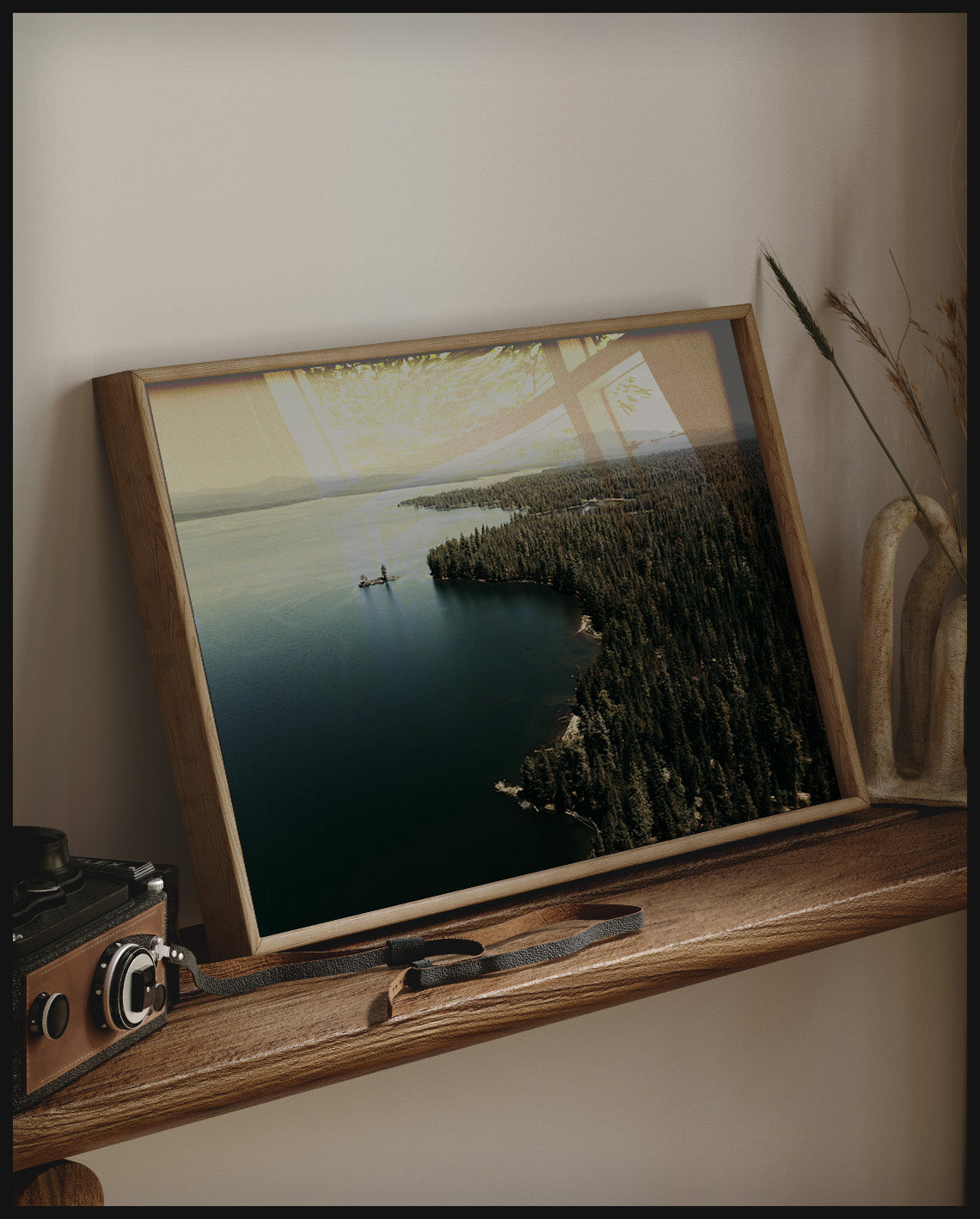 Ein gerahmtes Poster von einem See, umgeben von dichten Wäldern, aufgenommen aus der Vogelperspektive, auf einem Holzregal an die Wand angelehnt