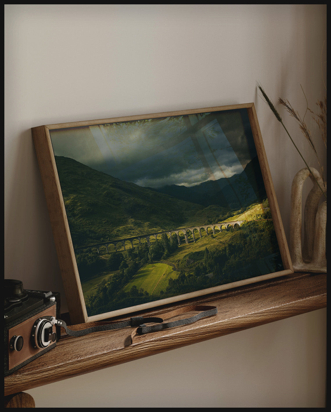 Ein gerahmtes Poster von dem Glenfinnan-Viadukt bei bewölktem Himmel, auf einem Holzregal an die Wand angelehnt