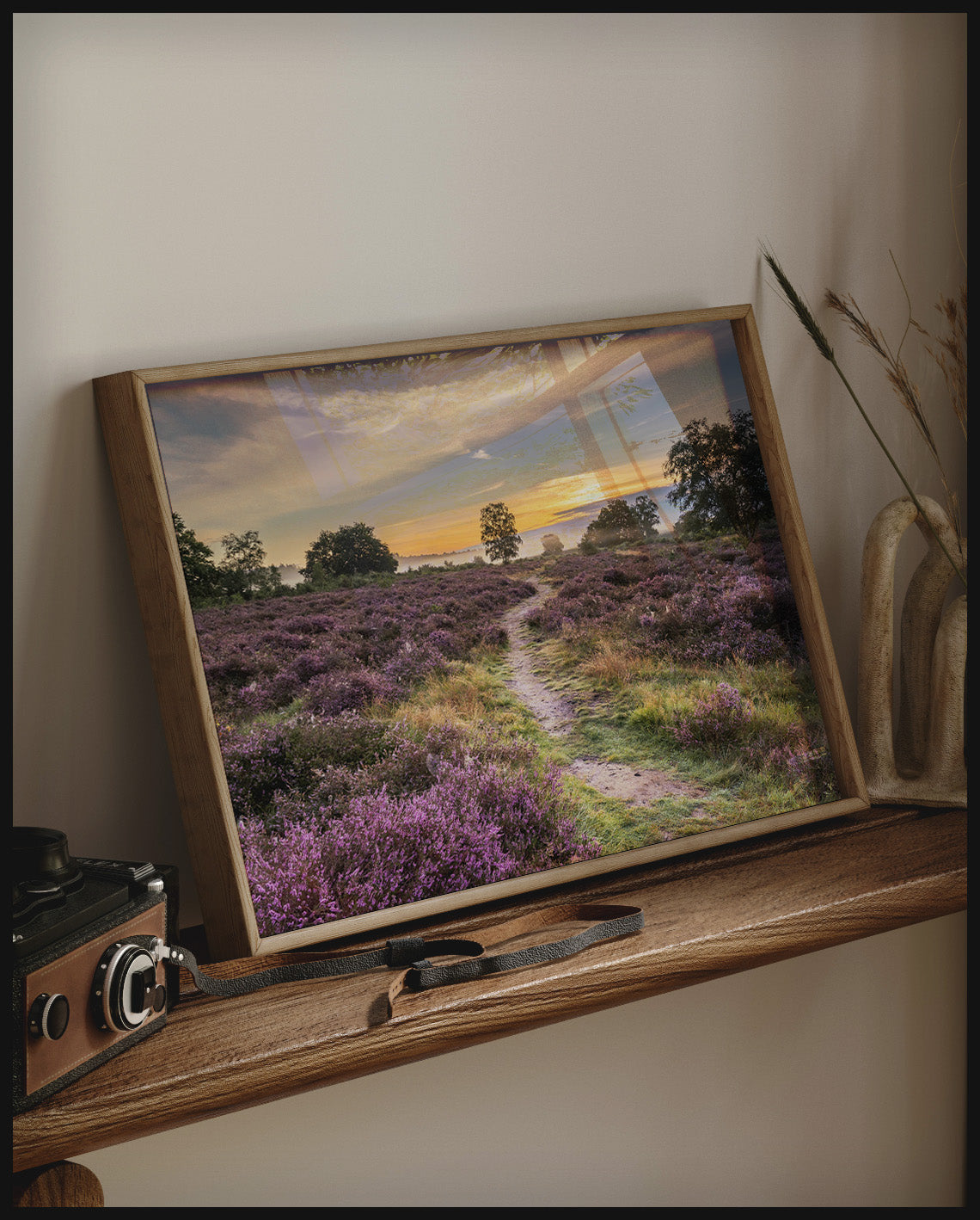 Ein gerahmtes Poster von einer Heidefläche mit lila Blumen im Sonnenuntergang, auf einem Holzregal an die Wand angelehnt