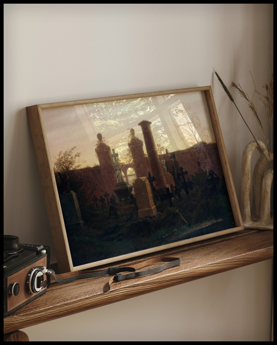 Ein gerahmter Kunstdruck von einem Friedhof mit einigen Gräbern und dem Sonnenuntergang im Hintergrund, auf einem Holzregal an die Wand angelehnt
