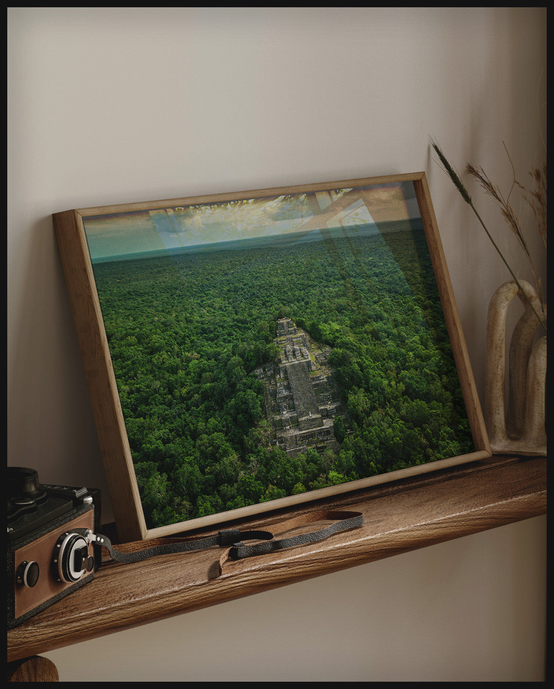 Ein gerahmtes Poster von einem antiken Maya-Tempel, der aus dichtem, grünem Dschungel herausragt, auf einem Holzregal an die Wand angelehnt