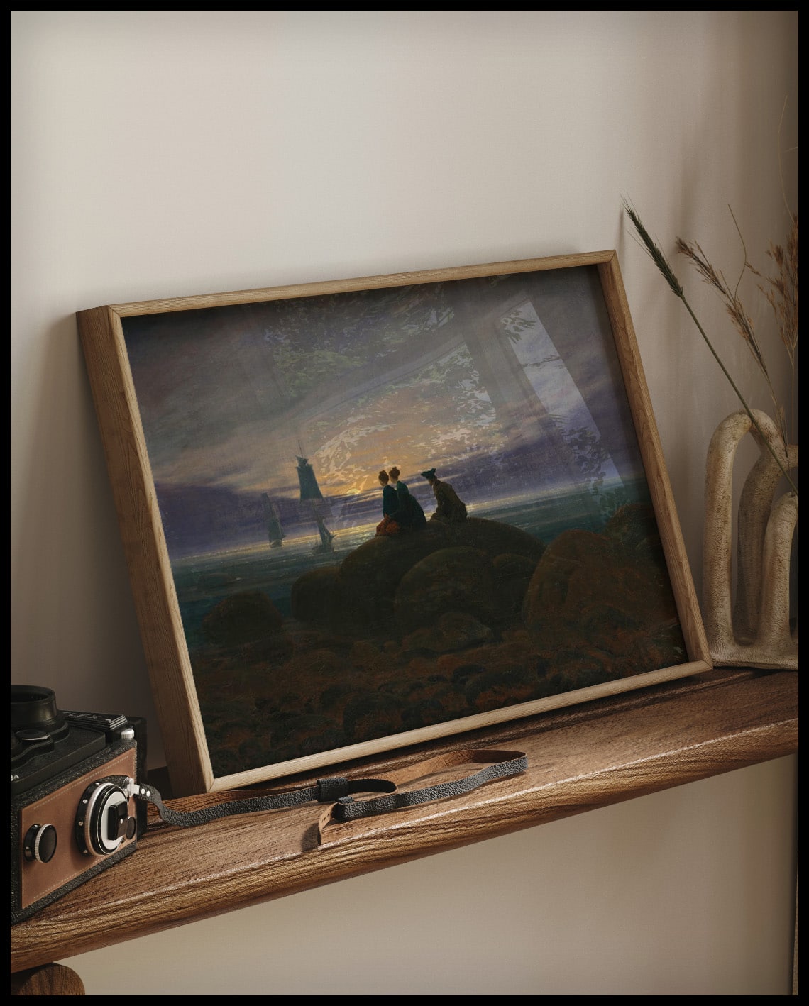 Ein gerahmter Kunstdruck von drei Personen am Meeresstrand die auf einem Felsen sitzen und den Mondaufgang betrachten, auf einem Holzregal an die Wand angelehnt