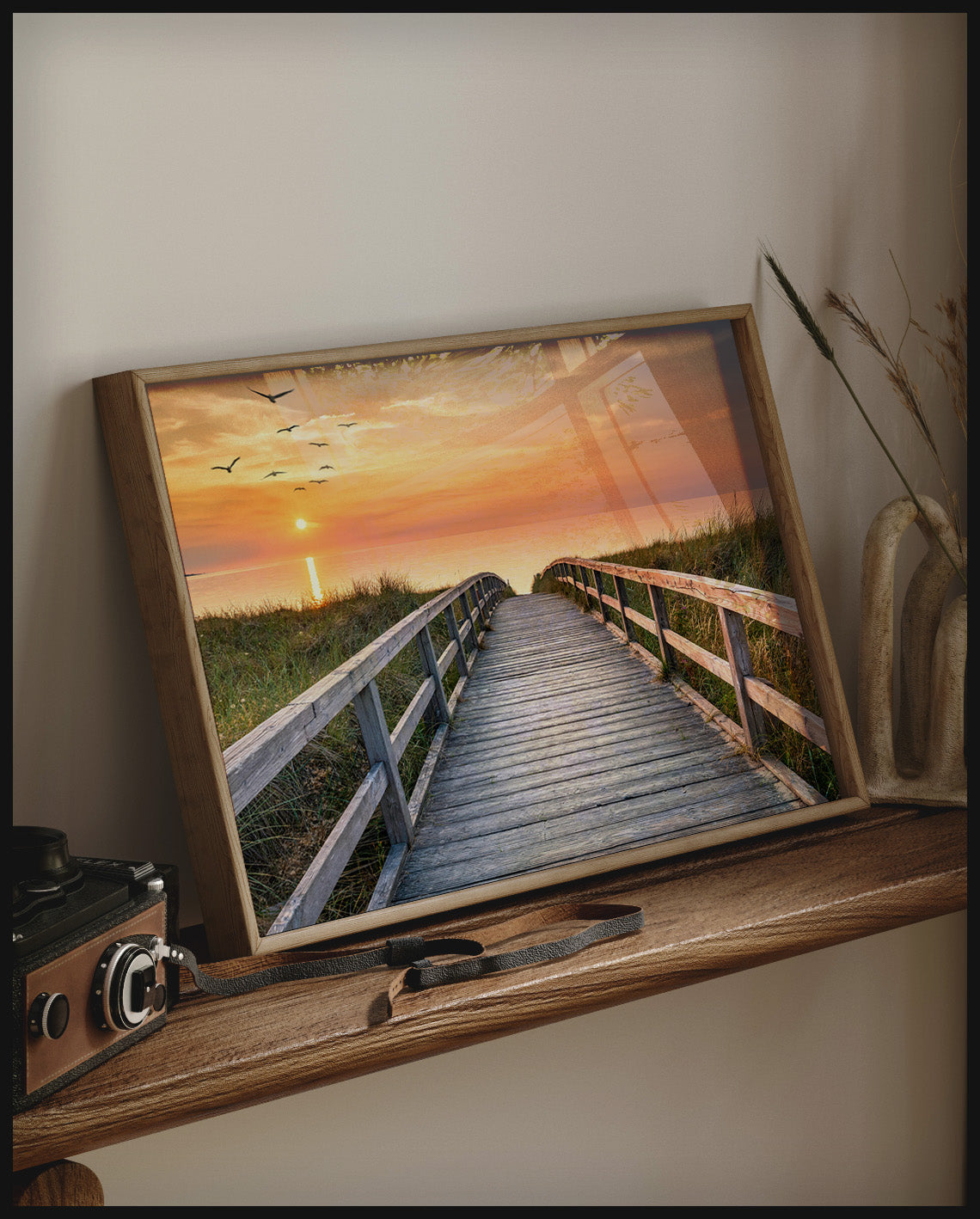 Ein gerahmtes Poster von einem Holzsteg im Sonnenuntergang, der über Dünen zum Strand führt, auf einem Holzregal an die Wand angelehnt