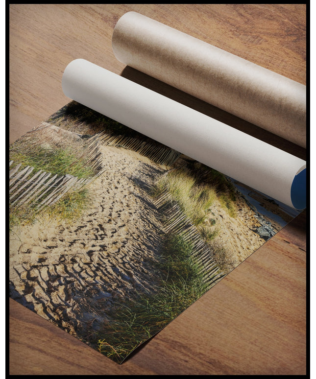 Ein Poster von einem sandigen Pfad, der sich entlang eines mit Gras und Dünen bewachsenen Küstenstreifens schlängelt, gerollt auf einer Holzoberfläche liegend
